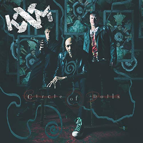 The three members of KXM sit before a dark background with framed doll images.