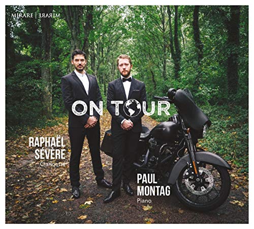 Two men in tuxedos with a motorcycle in a wooded area.