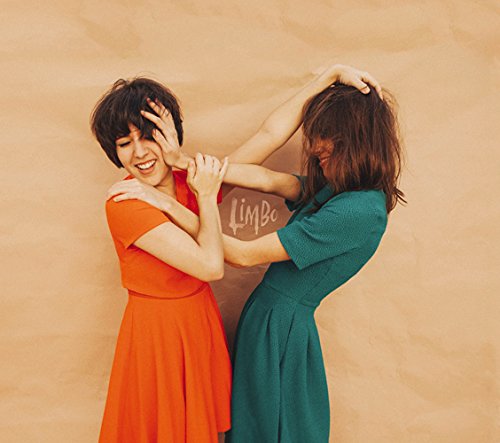 Two women in dresses playfully interact against a tan wall.
