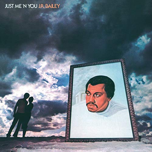 A couple silhouettes stand before a large framed portrait of J.R. Bailey under clouds.
