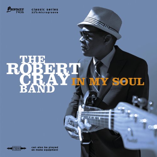 Robert Cray wearing a hat and suit while playing an electric guitar.