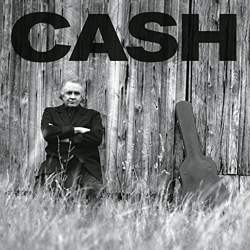 Johnny Cash sits with arms crossed before a wooden barn with a guitar case.