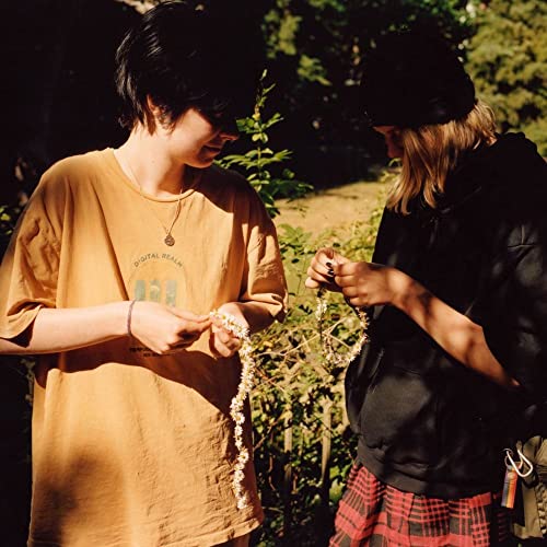 Two people outdoors, one braiding a garland of small white flowers.