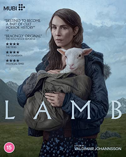A woman in a dark jacket holds a lamb in a rural landscape.