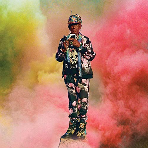 Lee Scratch Perry in patterned clothing, standing against a colorful, smoky background.