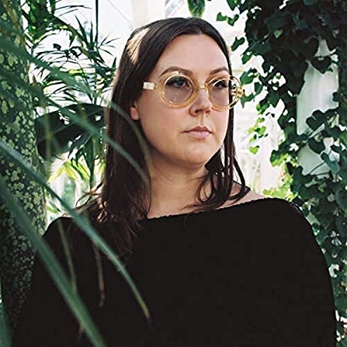 Nadia Reid portrait, wearing large round glasses, with foliage background.