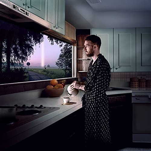 Man in patterned robe pours tea, looking out a kitchen window.