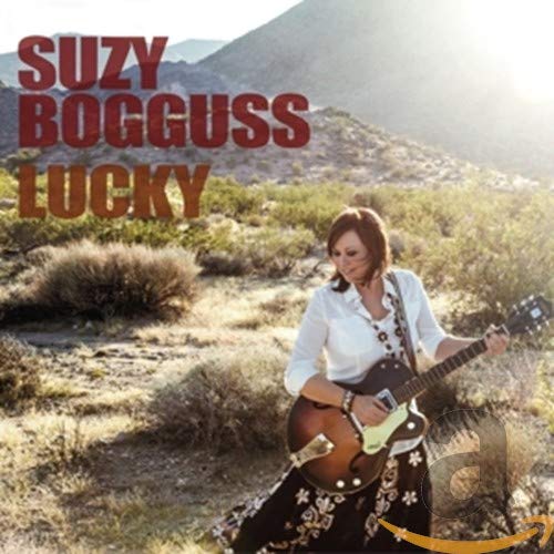 Suzy Bogguss playing an acoustic guitar in a desert landscape with mountains behind her.