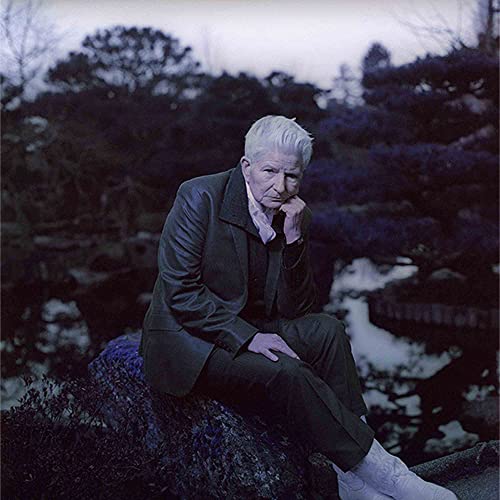 Older person with short white hair in a dark suit, outdoors.