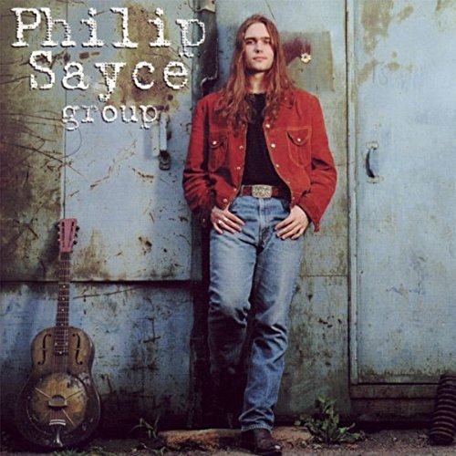 Long-haired man in a red jacket leaning against a blue door with a resonator guitar.