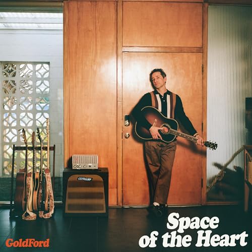 Man with guitar stands before a wooden door, with guitars and amp.
