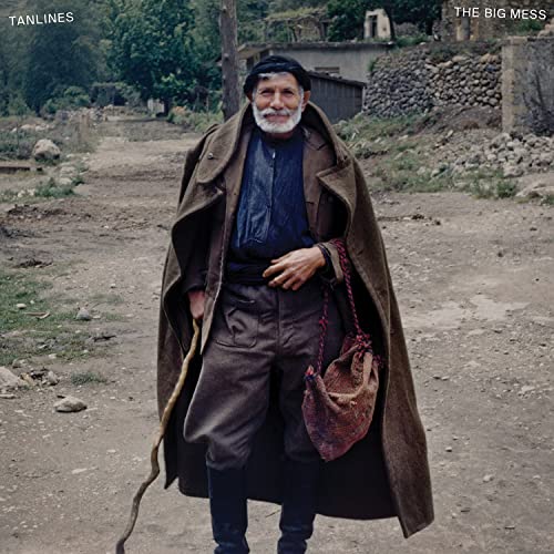 Older man in traditional clothing stands on a dirt road.