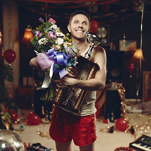 Jake Shears holds a trophy and flowers, smiling in a decorated room.