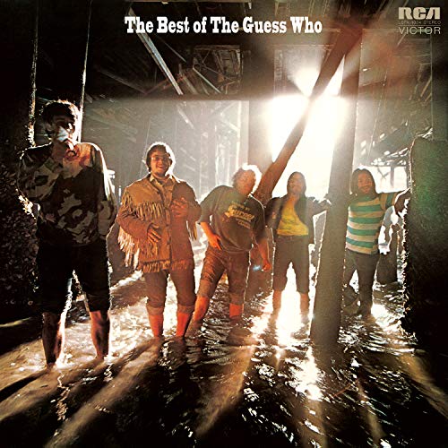 The Guess Who band members standing in shallow water under a pier at sunset.
