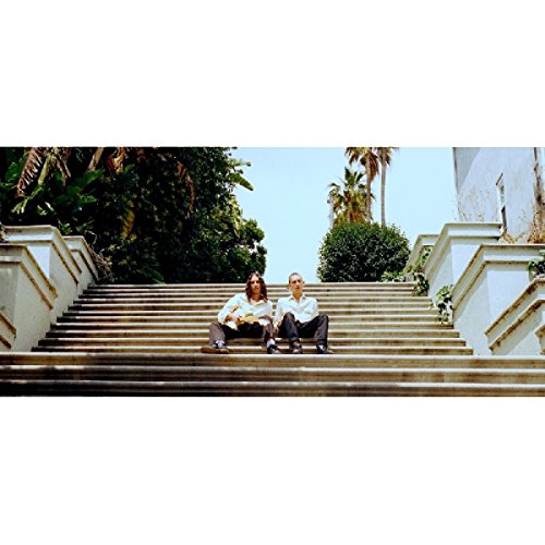 Two men sit on stone steps leading up to lush greenery.