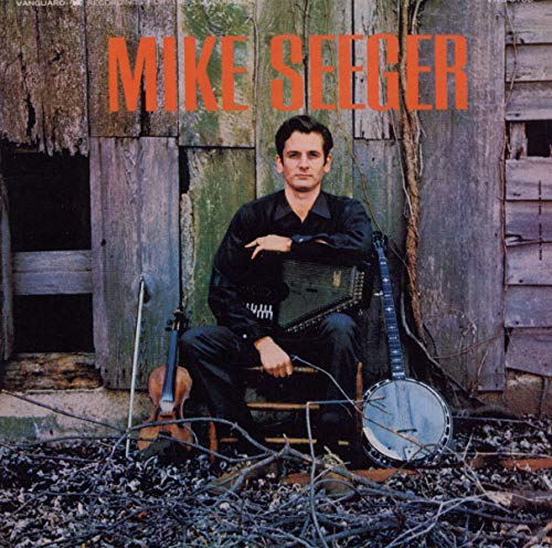 Mike Seeger sits with a banjo, fiddle, and accordion in front of a barn.