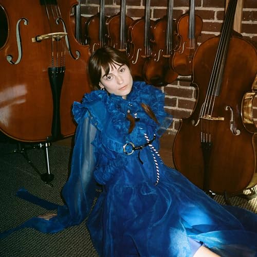 Woman in blue dress seated amongst several wooden stringed instruments.