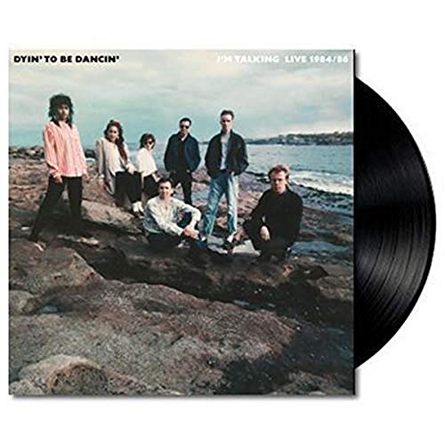 Six band members stand on a rocky beach with the ocean in the background.