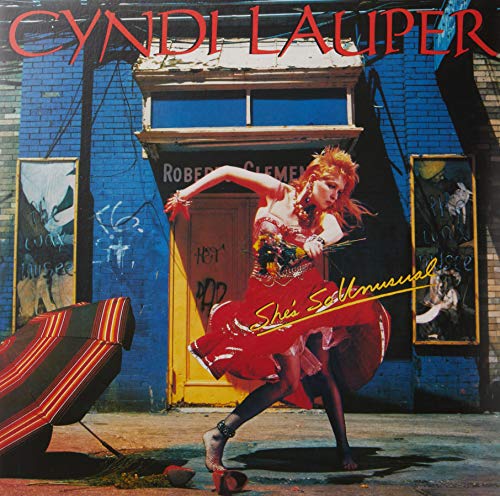 Cyndi Lauper in a red dress dancing in front of a blue brick building.