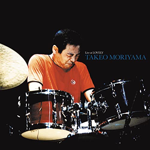 Takeo Moriyama playing drums on stage against a black background.