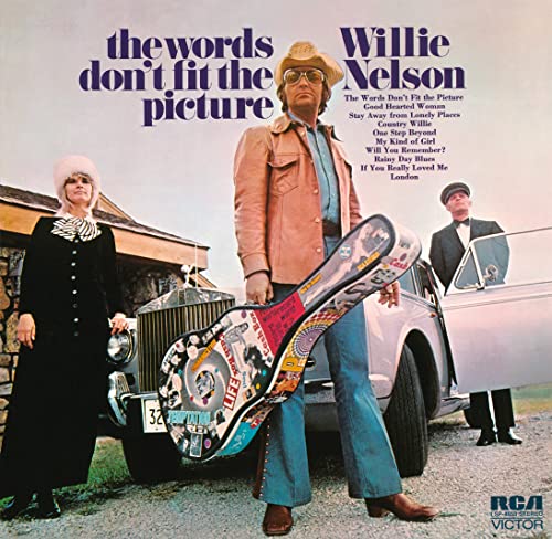 Willie Nelson holding a sticker-covered guitar case next to a car and two people.