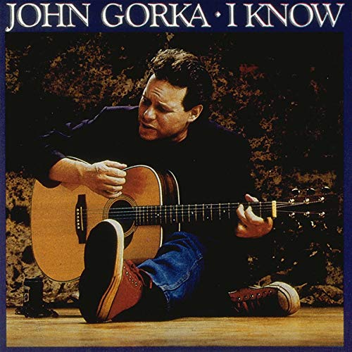 John Gorka sitting on the floor playing an acoustic guitar against a textured wall.