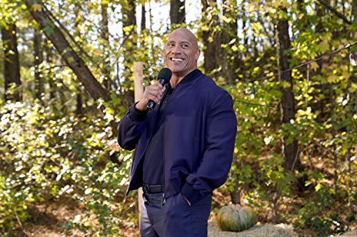 Dwayne Johnson wearing a navy jacket holds a microphone in a wooded outdoor setting.