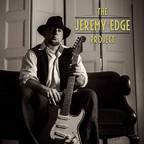 Man with guitar, wearing a hat, in front of a door.