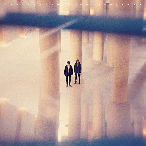 A man and woman stand between large pillars in a hazy, reflective architectural space.