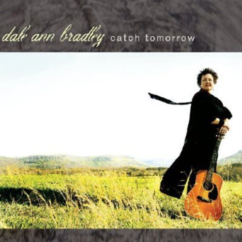 Dale Ann Bradley stands in a grassy field holding an acoustic guitar.