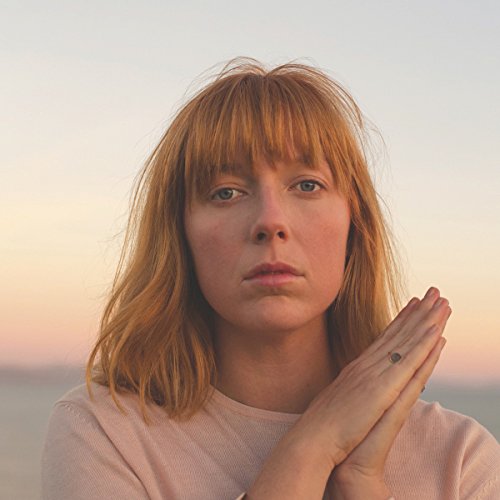Close-up portrait of a woman with reddish-blonde hair, hands clasped.