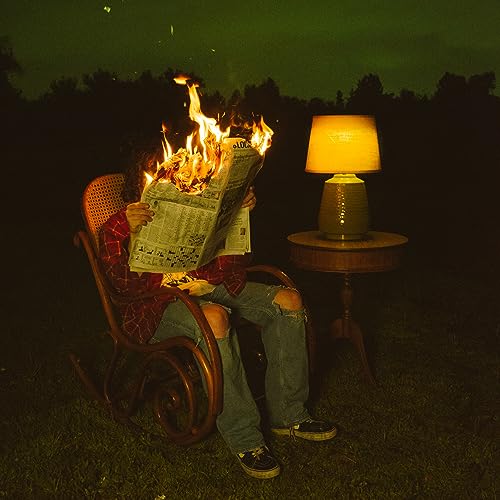 Person with flaming newspaper over face, seated with lamp nearby.