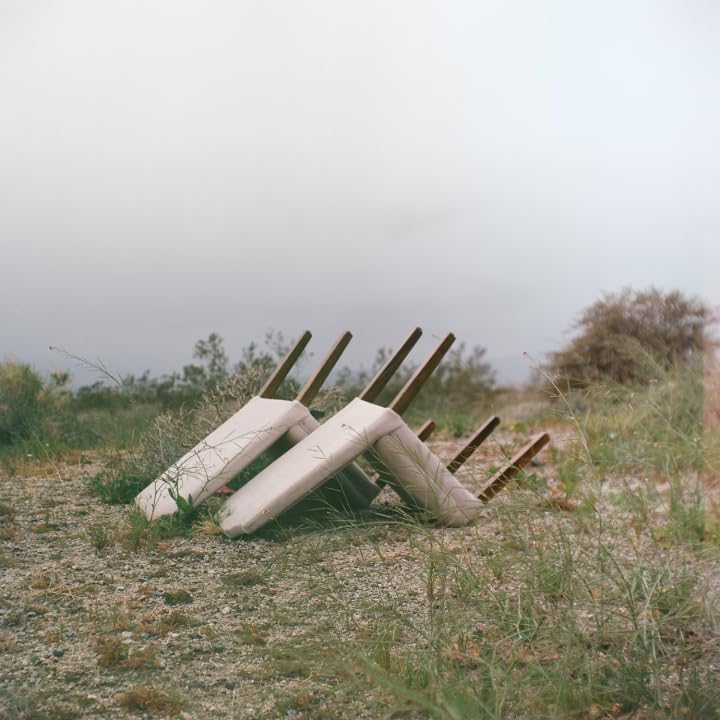 Stacked surfboards rest on a gravelly, overgrown hillside under a hazy sky.