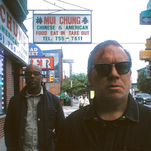 Two men in sunglasses stand on a city street with storefronts.
