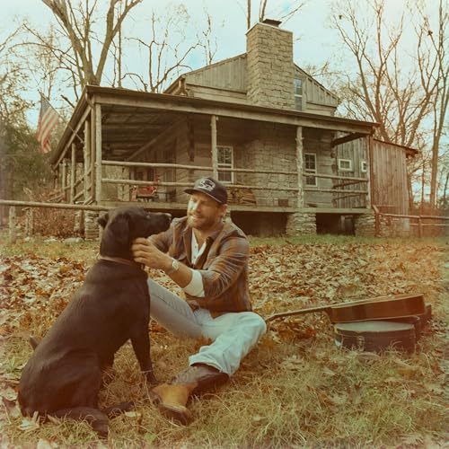 Chase Rice with a black Labrador in front of a rustic cabin.