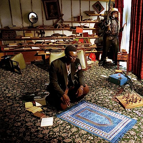 A man kneels on a patterned carpet before a prayer rug in a library.