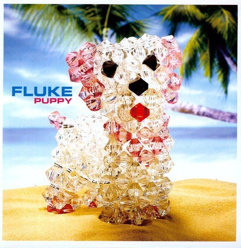 A crystal beaded puppy figurine stands on a sandy beach with palm trees behind.