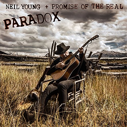 Neil Young with guitar, sitting outdoors against a cloudy sky.
