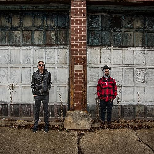 Two men in casual wear stand before a weathered brick and garage door facade.