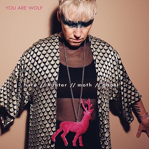 Person with dark eye makeup and a golden scale-patterned jacket, holding a red deer.