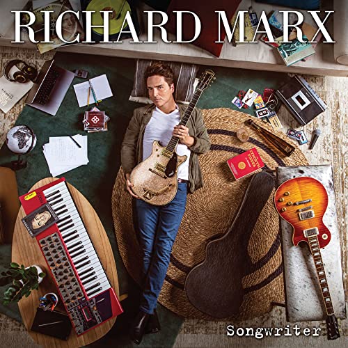 Richard Marx holding a guitar, surrounded by musical instruments and papers.