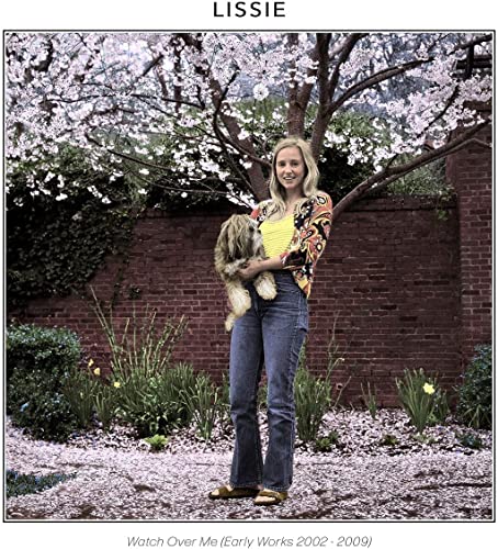 Lissie with a small dog, standing in front of a brick wall and blossoms.