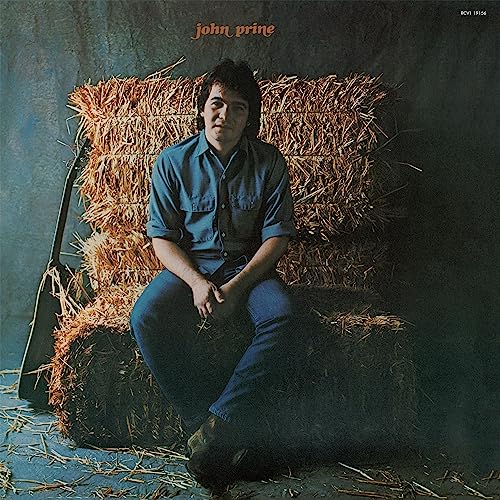 John Prine sitting on a hay bale with a guitar case leaning nearby.