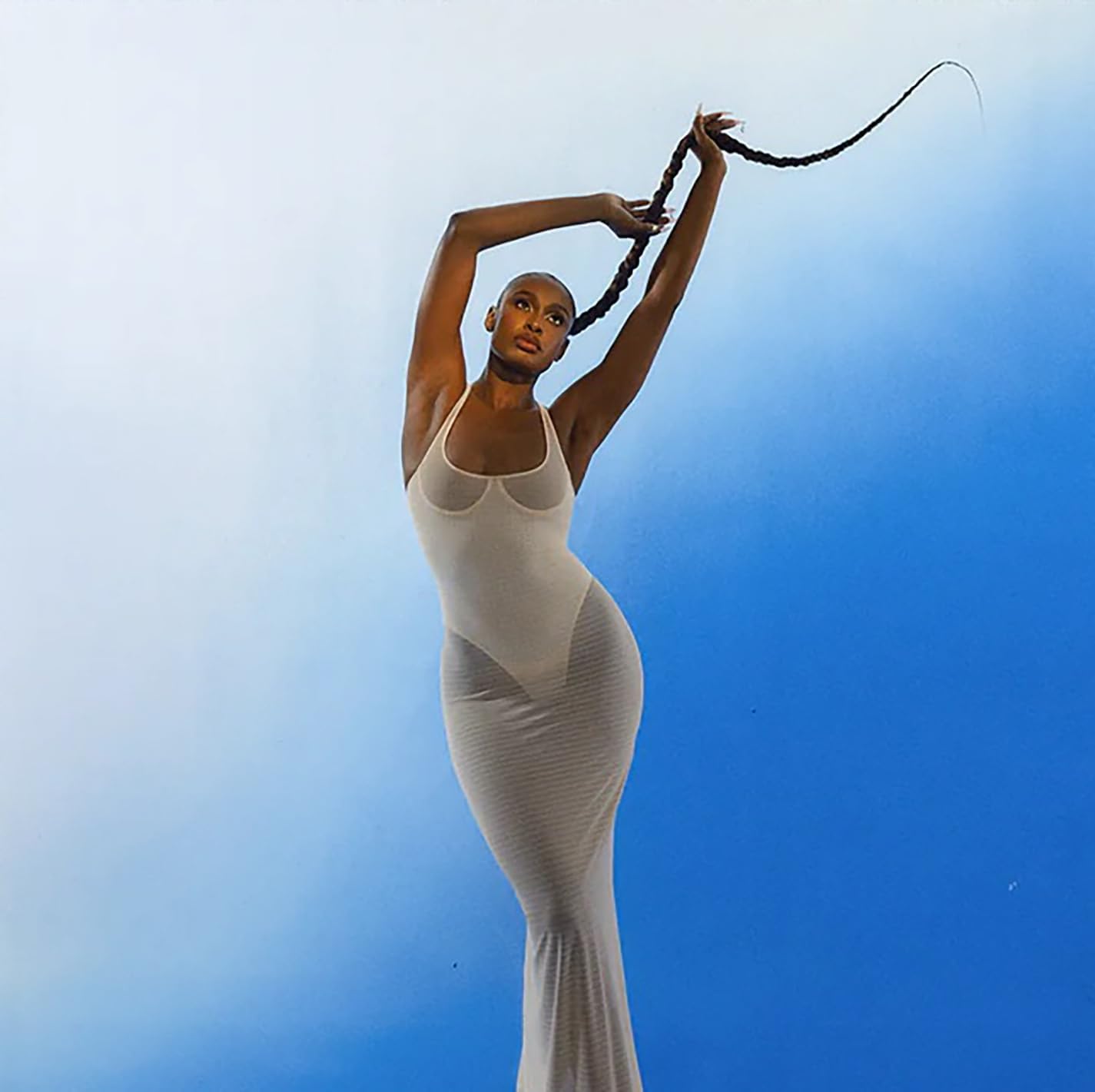 Coco Jones in a white dress, arms raised against a blue sky.