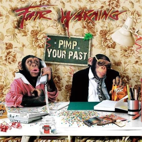 Two chimpanzees sit at a desk in front of a floral wallpaper background.