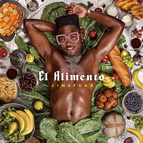 Cimafunk poses shirtless amidst a colorful spread of food and fruit.