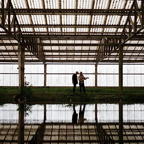Two figures with a guitar stand under a metal-roofed structure.
