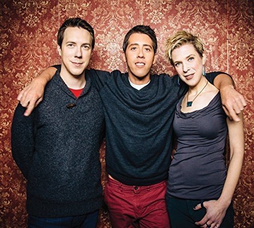 Three people smiling and embracing against a red patterned wall.