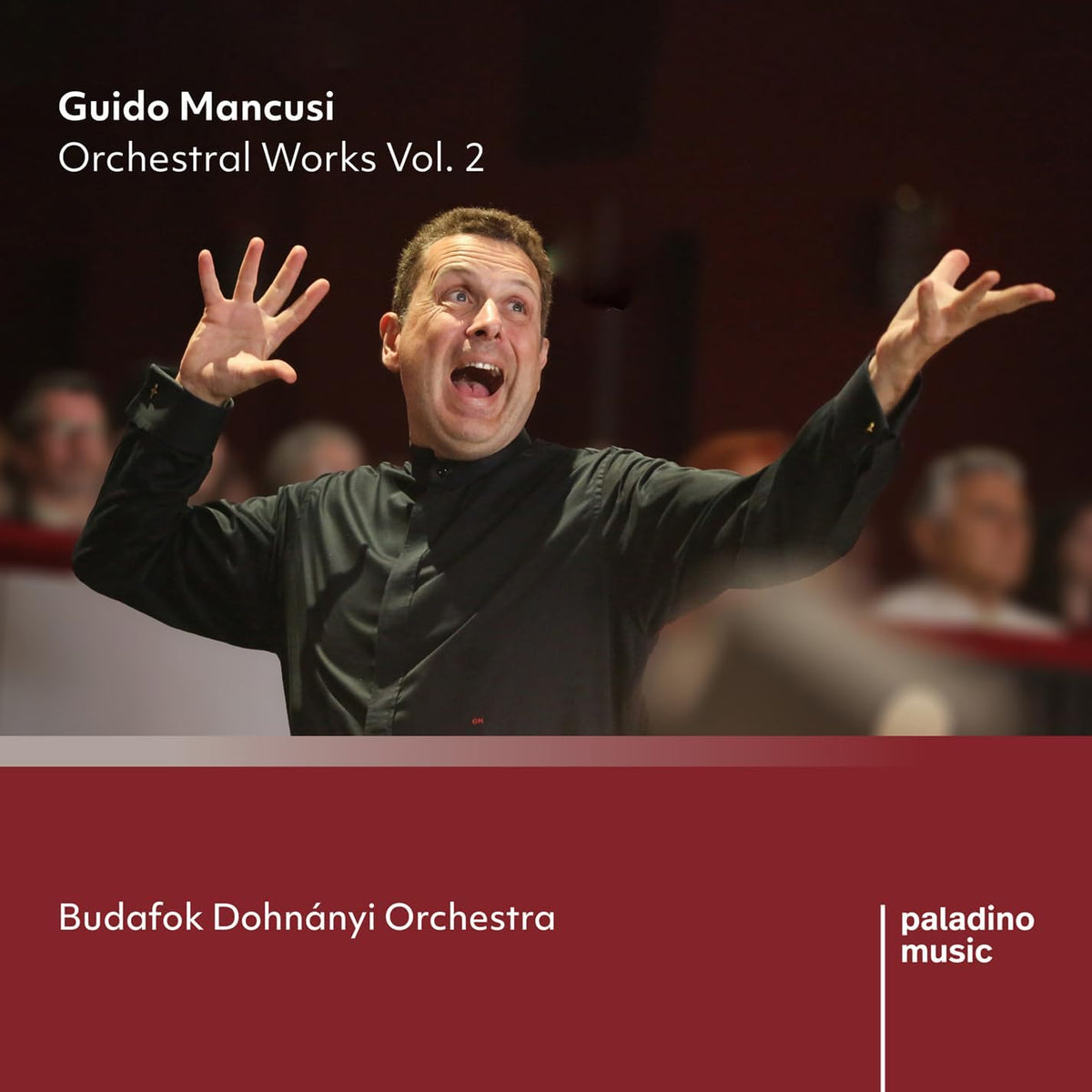 Conductor with arms raised, leading an orchestra in a red concert hall.