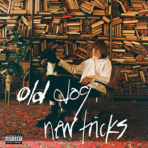 Man with a dog sits in a chair before a bookshelf.